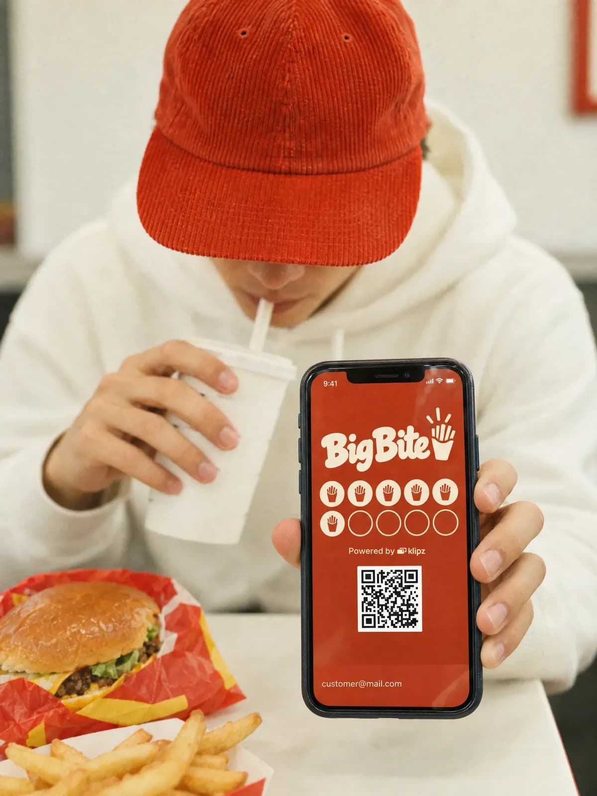En mand i rød kasket drikker af et papkrus på en burgerbar og holder telefonen frem med sit medlemskab hos Big Bite.