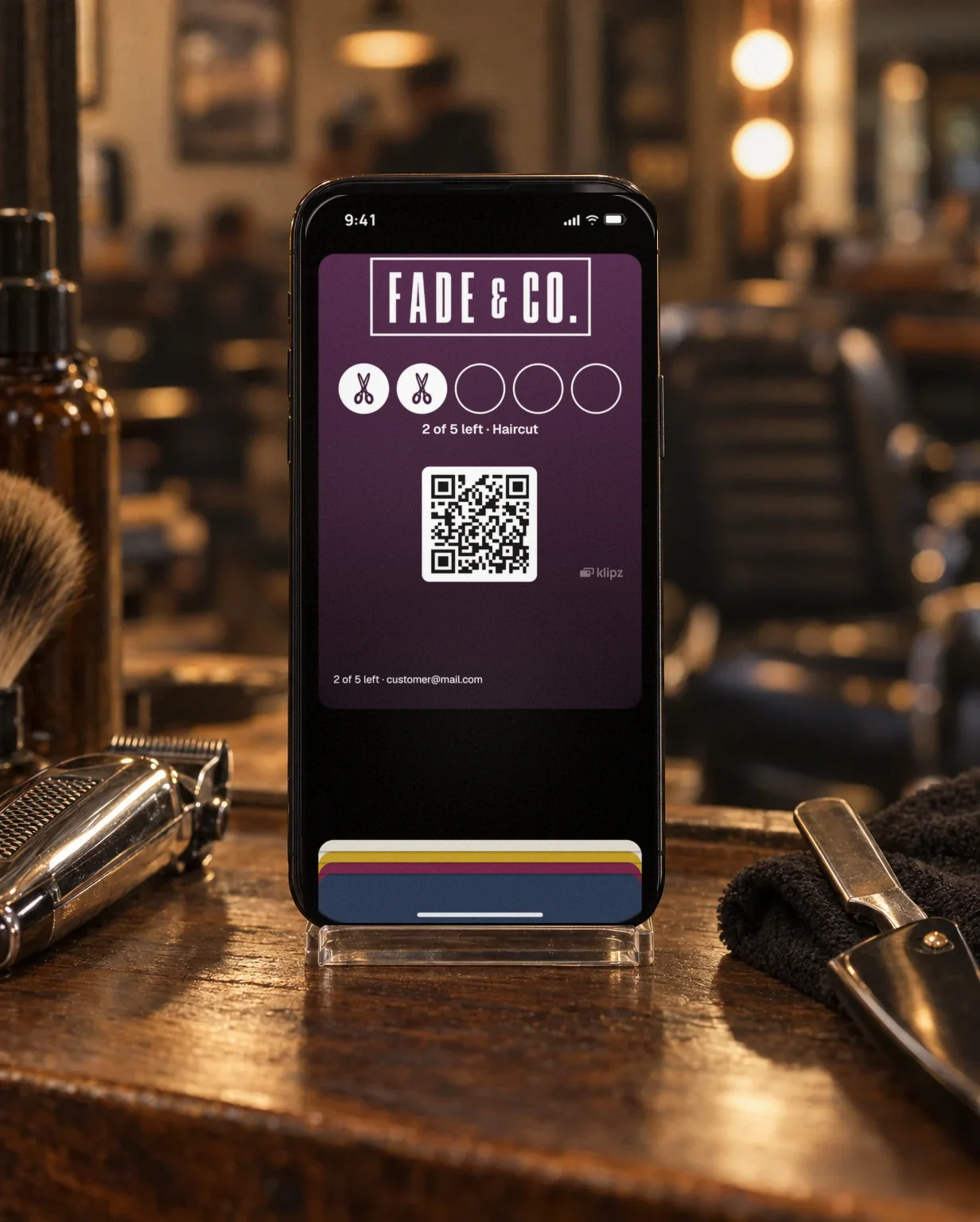 An iPhone in a clear stand on a wooden shelf in a barbershop by clippers and a straight razor. On the screen, Fade & Co.'s punch card is open in the wallet.
