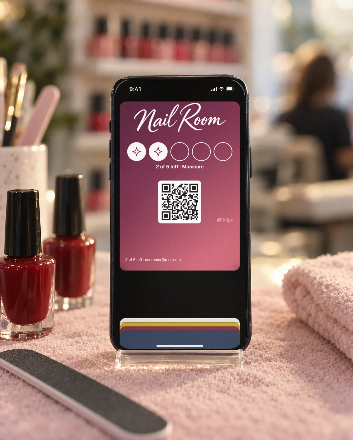 An iPhone in a clear stand on a pale pink towel by red nail polish and a nail file. On the screen, Nail Room's punch card is open in the wallet.