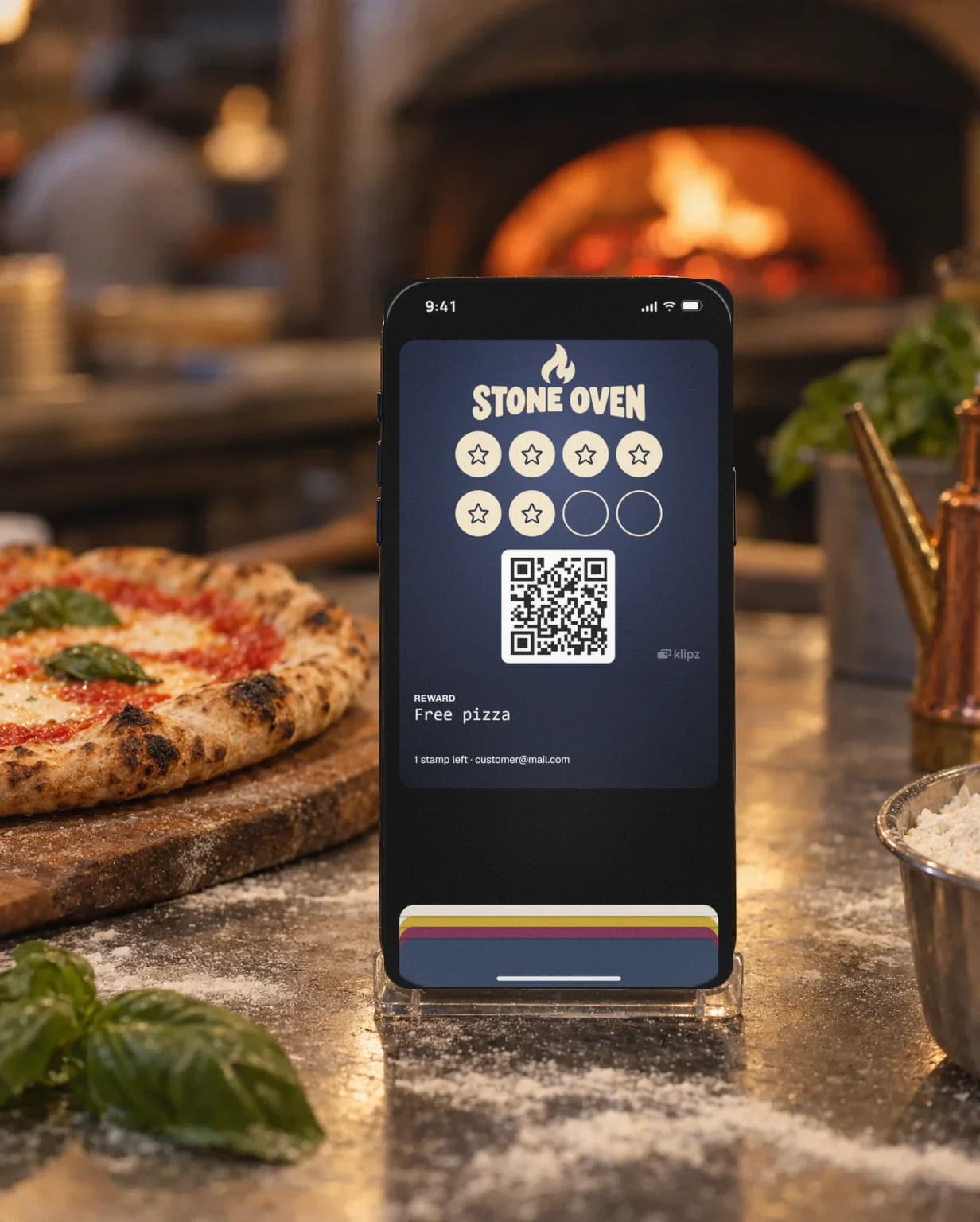 An iPhone in a clear stand on a pizzeria counter by a margherita on a wooden peel, a glowing pizza oven behind. On the screen, Stone Oven's stamp card is open in the wallet.