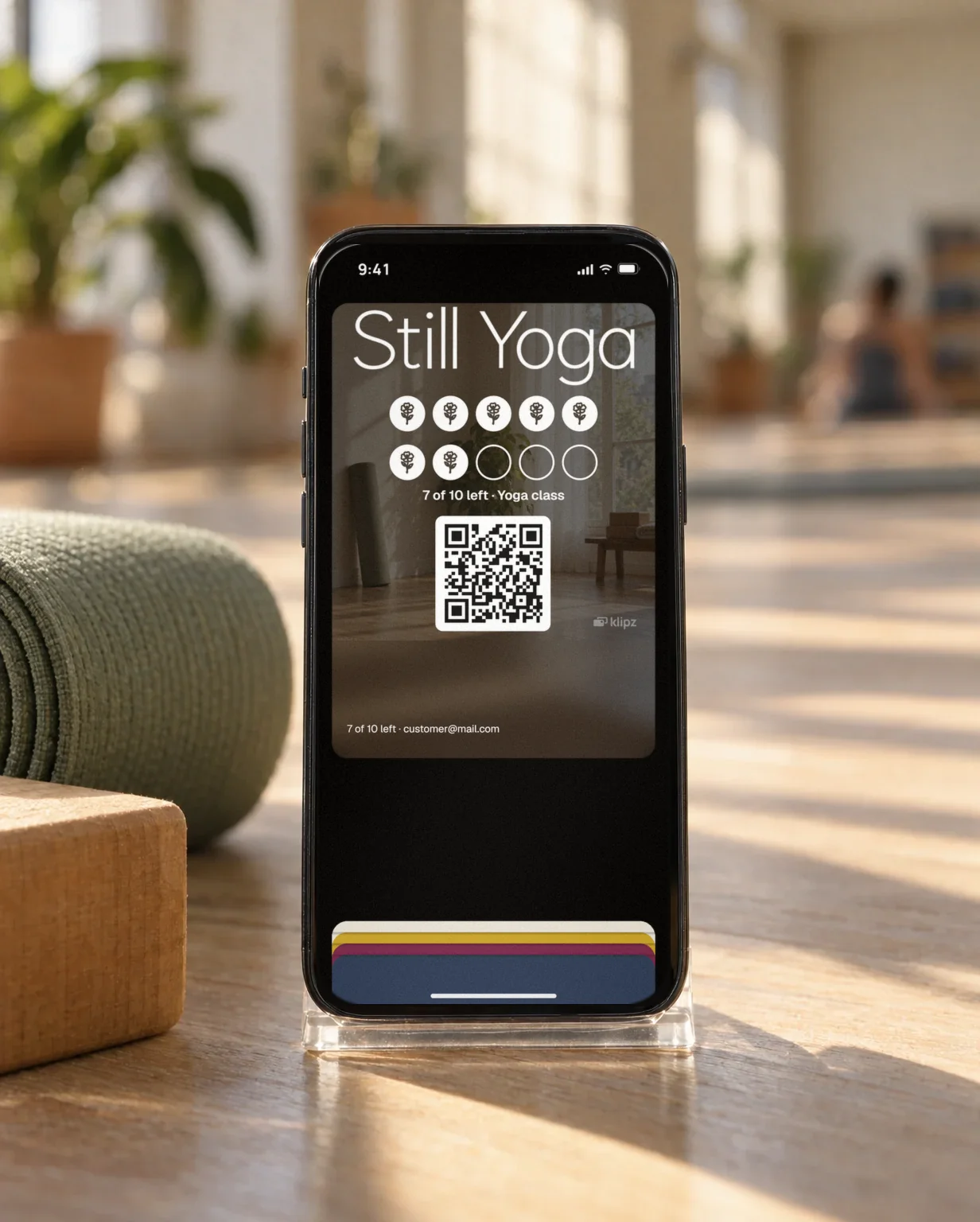 An iPhone in a clear stand on a pale wooden floor in a yoga studio by a rolled green mat and a yoga block. On the screen, Still Yoga's punch card is open in the wallet.